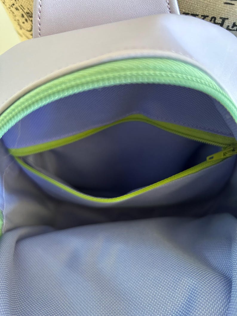 May include: A lavender-coloured bag with a green zipper and a light blue mesh lining. The bag has a small pocket on the inside.