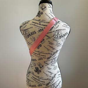 May include: A pink fabric strap is draped over a white fabric mannequin with a black stand. The mannequin is covered in a black and white print with the words "Paris 1889" repeated throughout.