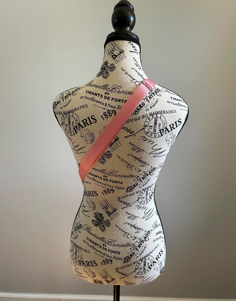 May include: A pink fabric strap is draped over a white fabric mannequin with a black stand. The mannequin is covered in a black and white print with the words "Paris 1889" repeated throughout.