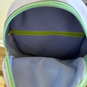 May include: A lavender-coloured backpack with a green zipper and a light green lining. The backpack is open and the inside is visible.