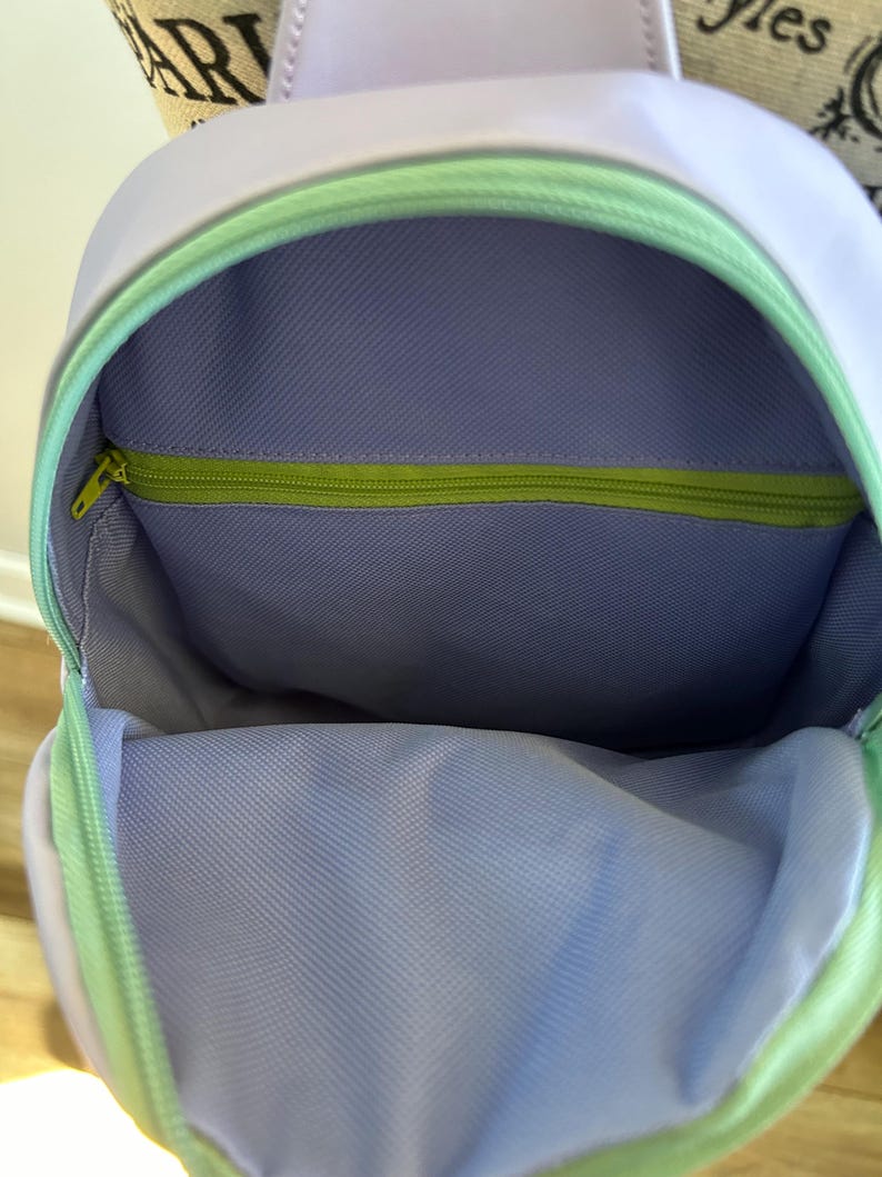 May include: A lavender-coloured backpack with a green zipper and a light green lining. The backpack is open and the inside is visible.