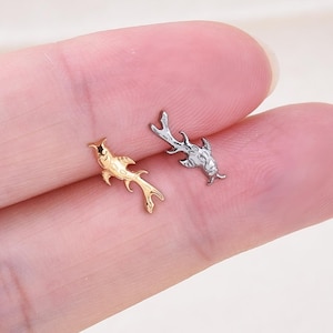 Op de afbeelding: Twee kleine, gedetailleerde oorbellen in de vorm van een vis. Eén is goudkleurig, de andere zilverkleurig. De oorbellen zijn op een lichtgekleurde achtergrond geplaatst. De oorbellen zijn waarschijnlijk van metaal.