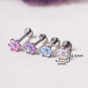 Puede incluir: Cuatro pendientes con forma de flor con pétalos iridiscentes en rosa, morado y azul. Los pendientes tienen un poste plateado y una parte trasera plana. Cada flor mide 5 mm de diámetro, con un poste de 4,5 mm. Se muestran sobre una superficie blanca.