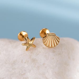 May include: A pair of gold-colored earrings, one shaped like a starfish and the other a seashell. The earrings are displayed on a white, textured surface against a light blue background. The starfish earring has five points, and the seashell earring has a detailed, ribbed design.