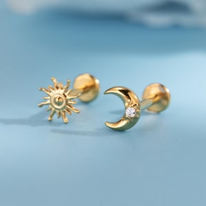 Könnte beinhalten: Goldfarbene Ohrstecker mit Sonnen- und Mond-Design. Der Sonnen-Ohrring hat detaillierte Strahlen, während der Mond-Ohrring einen kleinen, klaren Edelstein hat. Die Ohrringe sind vor einem weichen, blauen Hintergrund platziert.