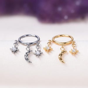 Op de afbeelding: Twee oorringen, één zilver en één goud, elk versierd met hemelse bedels. De zilveren oorring heeft een halve maan en twee sterren met heldere edelstenen. De gouden oorring heeft een maan met textuur en sterren met heldere edelstenen.