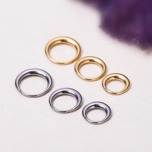 May include: An assortment of gold and silver-toned metal rings of varying sizes. The rings are arranged on a light-colored surface, with a blurred purple element in the background. The rings are likely jewelry components or findings.