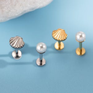 Puede incluir: Cuatro pendientes de botón sobre un fondo azul. Dos son plateados, uno con diseño de concha y otro con una perla. Los otros dos son dorados, uno con diseño de concha y otro con una perla.