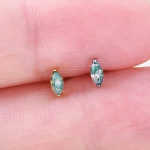 May include: A pair of small stud earrings. One earring has a gold post with a light green, oval-shaped stone. The other earring has a silver post with a similar light green, oval-shaped stone. The earrings are displayed on a person's finger.