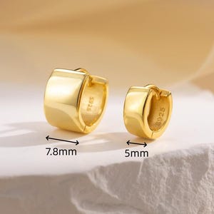 May include: Two gold-coloured hoop earrings. The larger earring measures 7.8mm, and the smaller one is 5mm. The earrings have a smooth, polished surface and a classic, minimalist design. The earrings are displayed on a white surface.