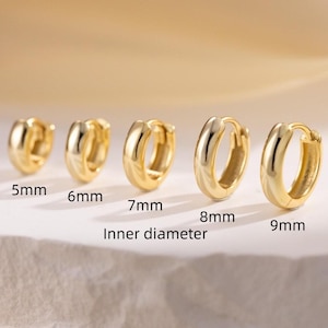 Puede incluir: Se muestran cinco pendientes de aro dorados de diferentes tamaños. Los diámetros interiores son de 5 mm, 6 mm, 7 mm, 8 mm y 9 mm. Los pendientes están dispuestos en fila sobre un fondo suave y borroso.