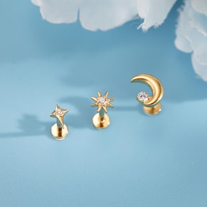 May include: Three gold-coloured stud earrings with celestial designs: a star, a sunburst, and a crescent moon. Each earring features a small, clear gemstone. The earrings are displayed on a light blue background.