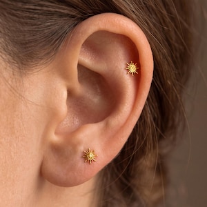 May include: Two small gold-toned sun-shaped stud earrings. One earring is positioned in the earlobe, and the other is placed in the upper cartilage area. Each stud features a central circular disc surrounded by thin, radiating sunburst rays.