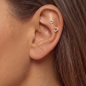 May include: Two gold-toned heart-shaped cartilage earrings featuring small, clear, sparkling stones set along the perimeter. The earrings are positioned in the upper ear, with one smaller heart placed above a slightly larger heart-shaped stud.
