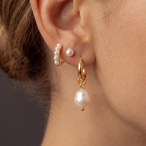 May include: A set of three gold-toned earrings consisting of a small hoop earring with five white pearls, a single white pearl stud earring, and a gold hoop earring featuring a dangling, irregular-shaped white baroque pearl charm.