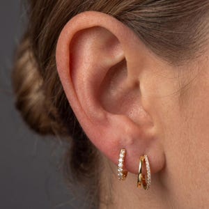 May include: Two gold hoop earrings worn in the earlobe. The left earring is a small hoop encrusted with clear stones. The right earring features a double-band design, with one smooth gold band and one band set with clear stones along the outer edge.