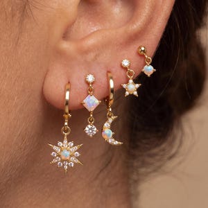 May include: A set of gold-toned earrings featuring celestial and geometric motifs. The collection includes a starburst charm, a diamond-shaped opal pendant, a crescent moon, and two small studs, all accented with white opal stones and clear crystals.