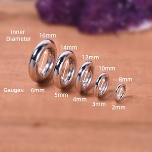 May include: A selection of silver-coloured, circular body jewellery, with inner diameters ranging from 2mm to 16mm. The gauges are labelled, and the jewellery is displayed on a wooden surface.