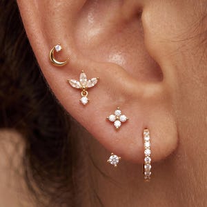 May include: A set of five gold earrings featuring a crescent moon stud with a clear stone, a marquise-cut floral cluster stud, a four-stone flower stud, a single round stone stud, and a small hoop earring encrusted with clear round stones.