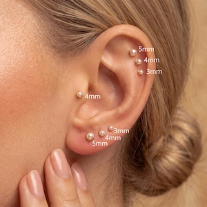 Puede incluir: Un conjunto de pendientes de botón dorados con perlas esféricas blancas de varios tamaños. La oreja muestra tres perlas a lo largo del cartílago superior de 3mm, 4mm y 5mm, una perla de 4mm en el trago y tres perlas en el lóbulo de 3mm, 4mm y 5mm.