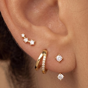 May include: A set of gold-tone earrings consisting of a three-stone curved stud, a double-banded hoop with a row of clear stones, and two individual round solitaire stud earrings, all featuring clear stones set in polished gold-tone metal.