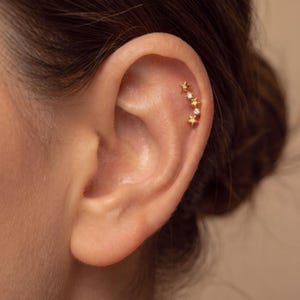 Puede incluir: Un pendiente de cartílago en tono dorado que presenta una fila curva de cinco estrellas pequeñas alternadas con dos piedras redondas transparentes. La pieza sigue la curva natural del cartílago superior de la oreja, creando un diseño delicado.