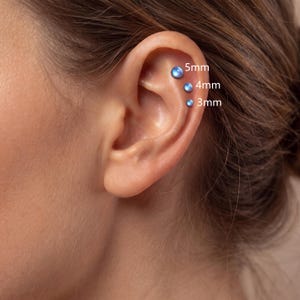 Könnte beinhalten: Drei runde, schillernd blaue Kugel-Ohrstecker sind vertikal entlang des Knorpels eines Ohrs angeordnet. Die Stecker sind mit ihren jeweiligen Größen beschriftet: 5mm für den oberen, 4mm für den mittleren und 3mm für den unteren Stecker.