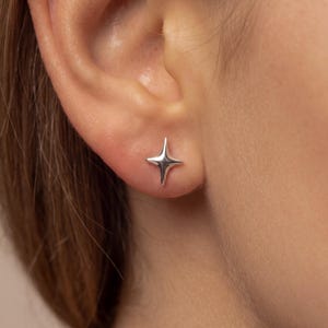May include: A small, polished silver stud earring shaped like a four-pointed star with elongated, tapered tips. The metal surface is smooth and reflective, catching the light to create subtle highlights across its curved, dimensional form.