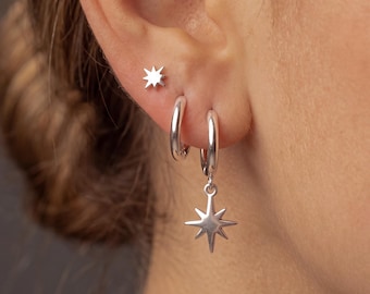 May include: A set of three silver-toned earrings consisting of one small star-shaped stud, one plain silver hoop, and one silver hoop featuring a dangling eight-pointed star charm. The jewelry pieces are polished and have a metallic finish.