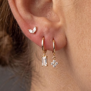 May include: A gold ear stack featuring a small stud with two marquise-cut clear stones, alongside two gold hoop earrings. One hoop holds a rectangular baguette-cut clear stone charm, and the other features a four-stone floral-shaped clear charm.