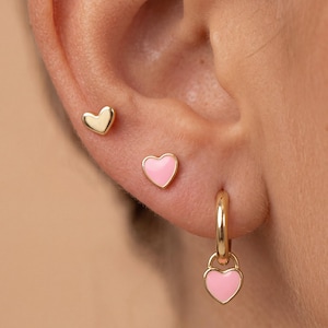 May include: A set of three gold-toned earrings consisting of one plain gold heart stud, one light pink enamel heart stud, and one gold hoop earring featuring a dangling light pink enamel heart charm, all worn in a single earlobe.