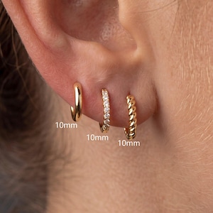 May include: Three gold hoop earrings, each measuring 10mm. The first is a smooth polished gold hoop, the second is a gold hoop set with small clear stones, and the third is a twisted gold rope-style hoop, all displayed in an earlobe.