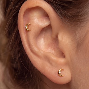 May include: Two gold-toned crescent moon earrings, each featuring a small, clear, round-cut gemstone set into the curve of the moon. One earring is positioned in the upper ear cartilage, while the other is placed in the lower earlobe.