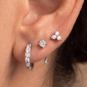 May include: A set of three silver-toned earrings featuring clear stones. One is a small hoop earring with four embedded stones, one is a single round stone stud, and the third is a cluster stud earring with three small round stones arranged together.