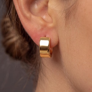 Peut inclure: Une petite créole épaisse en or poli avec une surface lisse et réfléchissante. La boucle d'oreille présente un design à large bande arrondie qui repose sur le lobe de l'oreille, mettant en valeur une finition jaune métallique brillante.