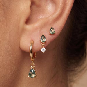 May include: A set of three gold-toned earrings featuring green moss agate stones. One earring is a small hoop with a dangling pear-shaped moss agate charm. The other two are stud earrings, one with a pear-shaped moss agate and one with a dangling clear stone.