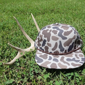 May include: A baseball cap featuring a brown, tan, and dark gray camouflage pattern with a flat brim and a thin brown cord accent across the base of the crown, positioned next to a set of light-colored deer antlers with multiple points.