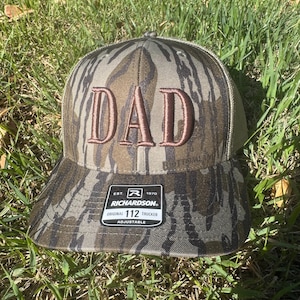 May include: Camouflage trucker hat with the word "DAD" embroidered in brown. The hat has a curved brim and a mesh back. The Richardson 112 Trucker hat is adjustable.