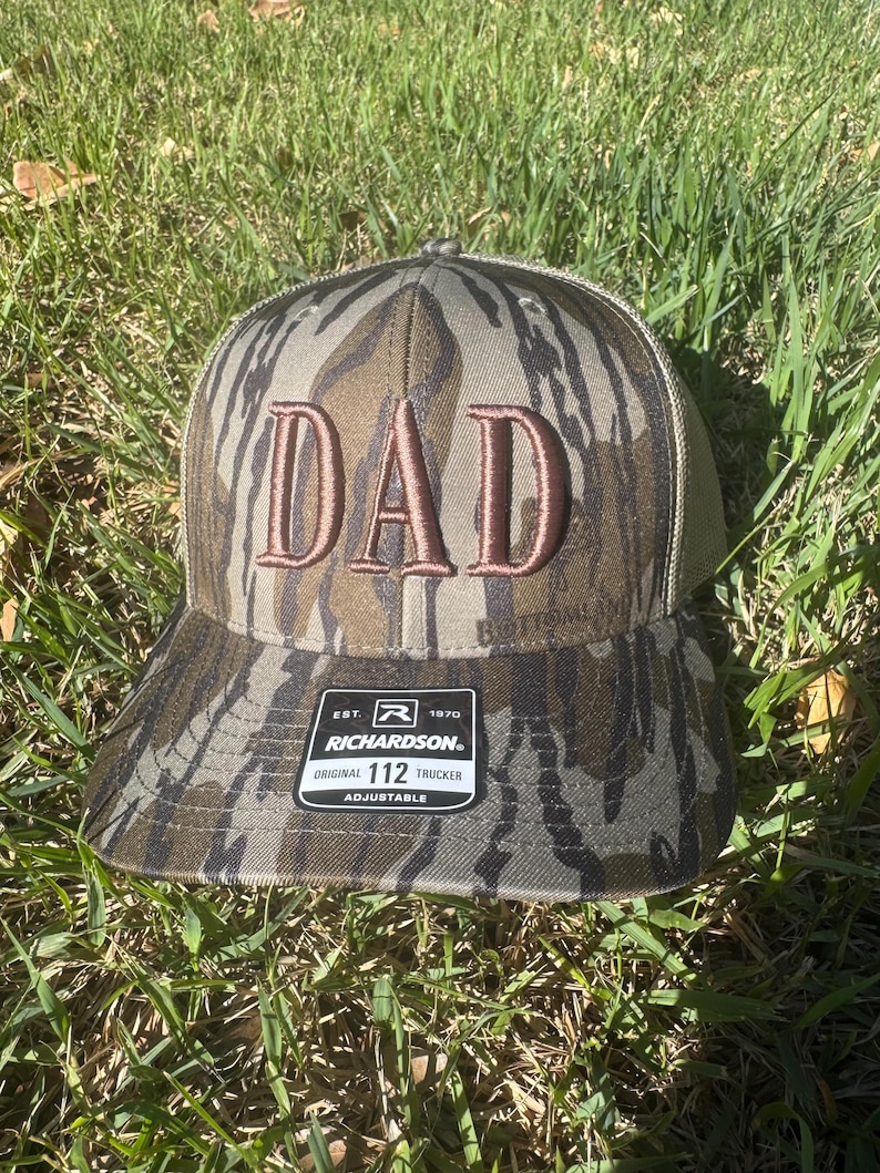May include: Camouflage trucker hat with the word "DAD" embroidered in brown. The hat has a curved brim and a mesh back. The Richardson 112 Trucker hat is adjustable.