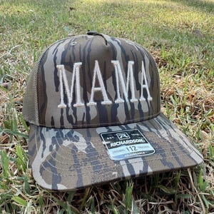 Op de afbeelding: Een camouflage trucker pet met het woord "MAMA" geborduurd in witte blokletters. De pet heeft een mesh achterkant en een gebogen klep. Een zwart-wit label is aan de klep bevestigd.