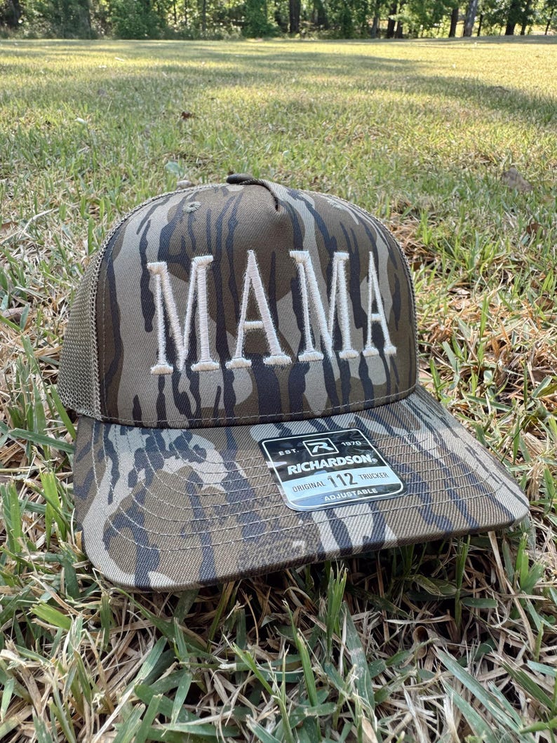 Op de afbeelding: Een camouflage trucker pet met het woord "MAMA" geborduurd in witte blokletters. De pet heeft een mesh achterkant en een gebogen klep. Een zwart-wit label is aan de klep bevestigd.