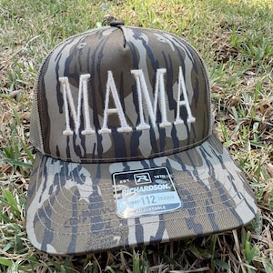 Op de afbeelding: Truckerpet met camouflagepatroon met het woord "MAMA" geborduurd in witte blokletters. De pet heeft een mesh achterkant en een gebogen klep. Een label op de klep luidt "Richardson 112 Trucker Adjustable".