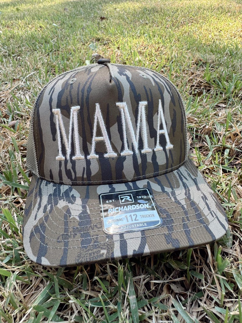 Op de afbeelding: Truckerpet met camouflagepatroon met het woord "MAMA" geborduurd in witte blokletters. De pet heeft een mesh achterkant en een gebogen klep. Een label op de klep luidt "Richardson 112 Trucker Adjustable".