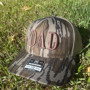 May include: A camouflage trucker hat with the word "DAD" embroidered in a brown color. The hat has a mesh back and an adjustable strap. The Richardson brand label is visible.