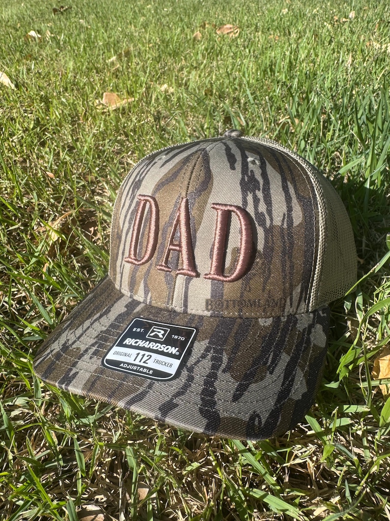 May include: A camouflage trucker hat with the word "DAD" embroidered in a brown color. The hat has a mesh back and an adjustable strap. The Richardson brand label is visible.