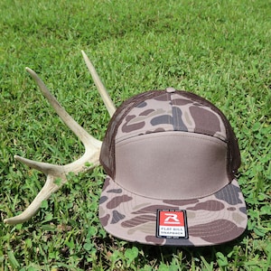 May include: A camouflage print snapback cap with a solid brown front panel and brown mesh back. The hat features a flat bill with a black and red sticker that reads Richardson Flat Bill Snapback. A set of light-colored deer antlers sits behind the cap.