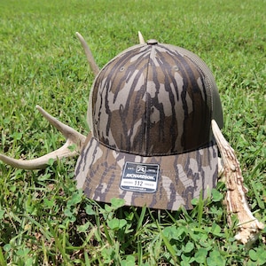 May include: A brown and tan camouflage trucker hat with a mesh back and a structured front panel. The front features a black rectangular sticker with the text Richardson 112 Trucker. The hat is positioned between two light-colored deer antlers.