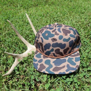 May include: A baseball cap featuring a brown and dark blue camouflage pattern with a brown cord accent across the base of the crown, positioned next to a light-colored deer antler. The cap has a structured crown and a flat brim.