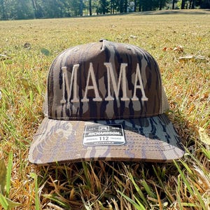 Op de afbeelding: Een camouflage trucker pet met het woord "MAMA" geborduurd in witte blokletters. De pet heeft een gebogen klep en een mesh achterkant. De Richardson 112 pet is verstelbaar.