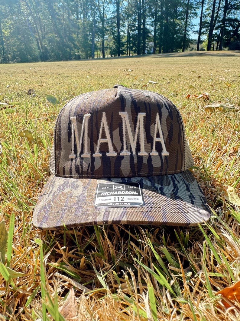Op de afbeelding: Een camouflage trucker pet met het woord "MAMA" geborduurd in witte blokletters. De pet heeft een gebogen klep en een mesh achterkant. De Richardson 112 pet is verstelbaar.
